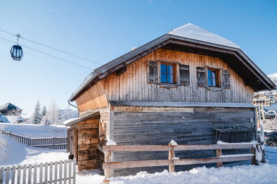 Almchalet aus Altholz im Schnee, dahinter die Gondelbahn