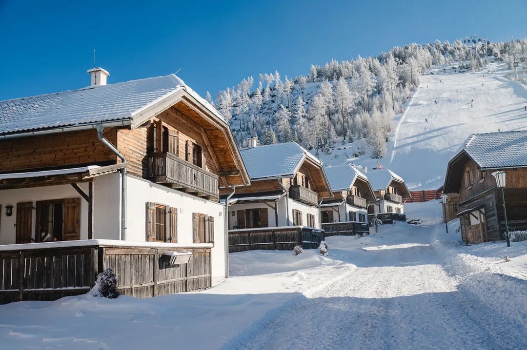 Verschneite Almhütten an der Dorfstraße, dahinter die Skipiste